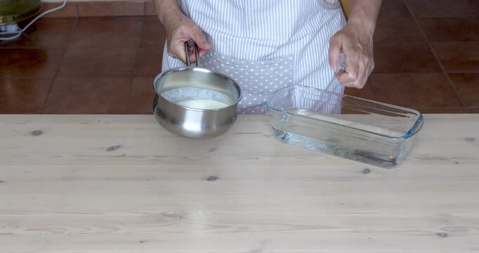 Softened Gelatin Squeezed and Added to Warm Cream in a Saucepan for Cheesecake. Close-up of female hands squeezing softened gelatin sheets and placing them into a saucepan containing warm liquid cream
