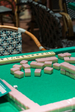 Mahjong Tiles on Green Felt: A close-up view of pink Mahjong tiles scattered across a green felt table, set against a backdrop of wicker chairs, hinting at an engaging game ready to begin.