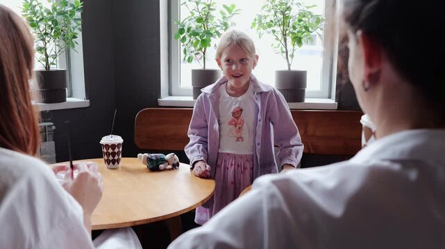 Young girl standing between two round wooden tables in cafe while adults converse and child shifts from folded arms to engaged smile then returns to attentive pose