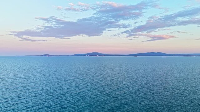 The ocean reflects shades of blue and pink as the sun sets in the background. Distant hills outline the landscape under a sky filled with clouds.