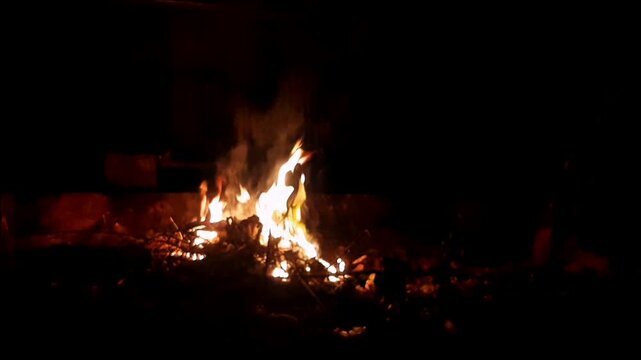 A small campfire burning brightly at night, surrounded by dry wood and natural ground. The warm glowing flames contrast beautifully with the dark background, creating a cozy atmosphere associated with