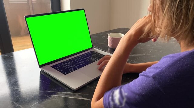 Woman using laptop in over-the-shoulder view at home with green screen display, suited for remote work, online learning, website mockup, app presentation and digital advertising content replacement