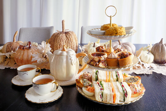 Autumn theme afternoon tea table scene with pumpkin spice tea, scones, cozy pumpkin, pecan and apple desserts and fall theme finger sandwiches and savory items