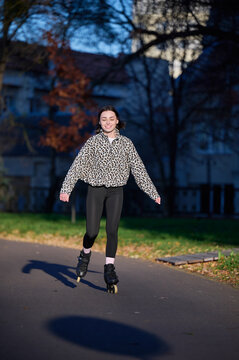 Young woman roller skating in park and smiling at camera, full length on paved path in evening light