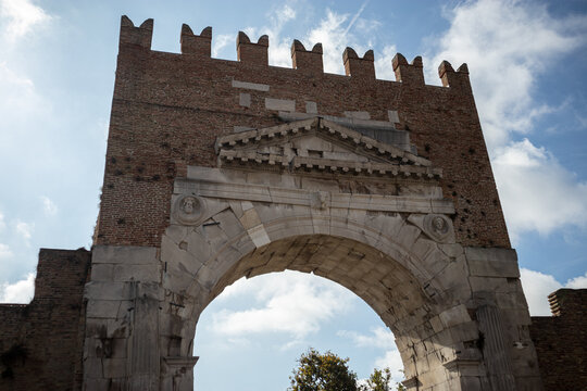 Prospettiva frontale dell'antico Arco d'Augusto a Rimini con la sua monumentale struttura in pietra bianca, capitelli corinzi e merlatura medievale superiore incorniciata da un cielo azzurro intenso