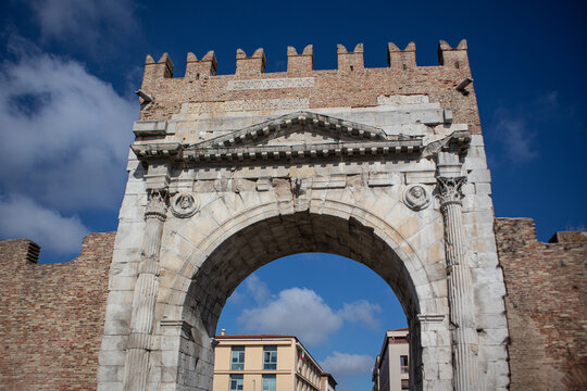 Prospettiva frontale dell'antico Arco d'Augusto a Rimini con la sua monumentale struttura in pietra bianca, capitelli corinzi e merlatura medievale superiore incorniciata da un cielo azzurro intenso