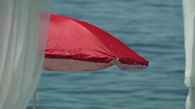 Bright red parasol flutters over turquoise ocean water at a seaside resort terrace. Static medium telephoto shot.