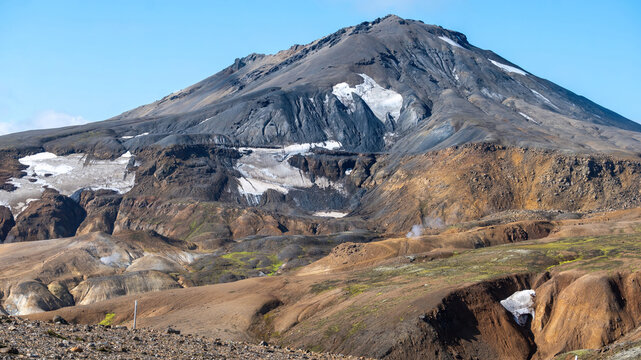 A dark volcanic peak with lingering snow rises above steaming geothermal vents and colorful moss-covered slopes near Kerlingarfjoll, Icelandic highlands.