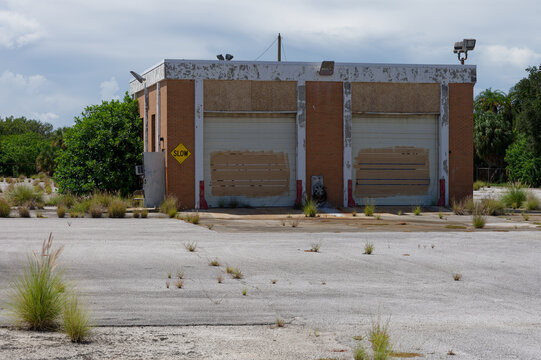 An abandoned empty parking lot with cracked asphalt, tire marks and overgrown weeds in front of a shuttered industrial building behind a chain-link fence under cloudy skies, evoking urban decay and ne