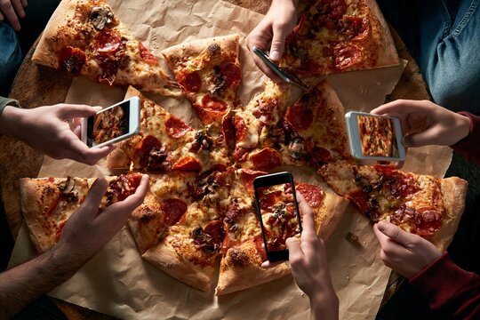 Group of friends using smartphones to photograph a large pepperoni pizza on a rustic paper surface