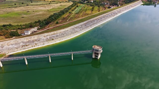 A large water reservoir contains a control tower at its edge. Surrounding areas include fields and trees, showing a mix of nature and man-made structures. The image captures a clear view of the scene.