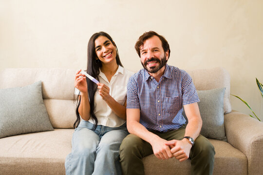 Happy couple sitting on sofa at home, feeling excited and looking at positive pregnancy test result