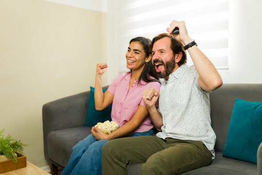 Pretty couple watching television, cheering with excitement, holding popcorn bowl during a fun movie night at home