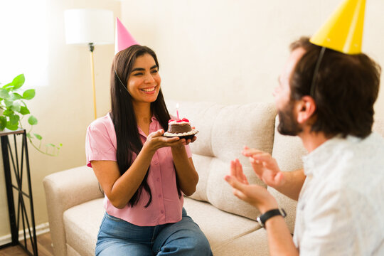 Young couple celebrating birthday at home, woman smiling and offering a small cake with candle to her partner