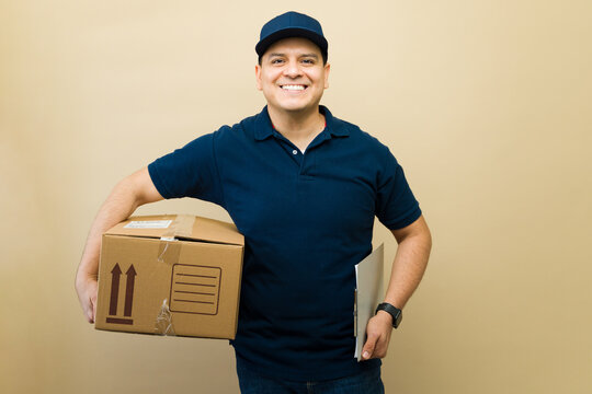 Hispanic delivery man wearing uniform cap holding package and clipboard providing shipping service