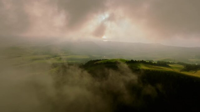 Aerial view of rolling green hills on Sao Miguel Island, partially covered by thick mist and dramatic clouds during a soft, atmospheric sunset.