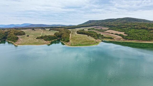 A wide water body reflecting the cloudy sky, surrounded by green hills and open fields. The landscape has patches of trees and clear areas of grass.