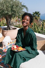 Obraz premium Smiling woman in a green dress holding a plate of fresh fruit on a terrace garden with cat and ocean view for summer lifestyle banner and wellness design