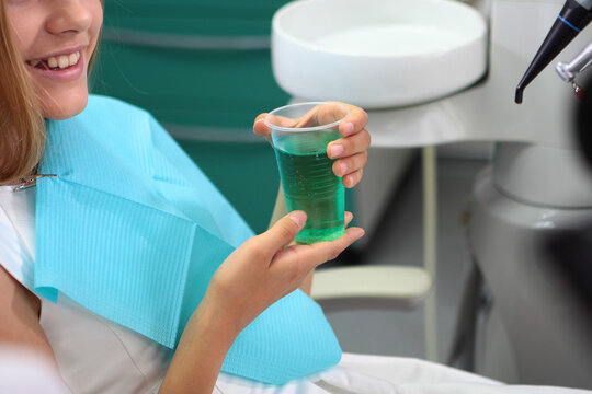 Girl holds green cup of mouthwash in dentist's chair. She wears blue bib and white uniform. Smiling patient rinses her mouth. Professional oral hygiene procedure