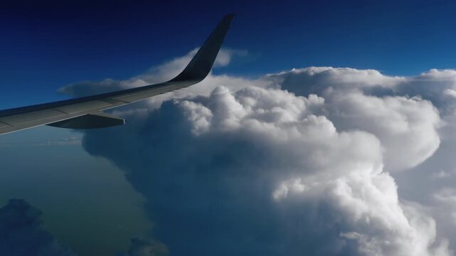 Airplane wing flying high above massive white cumulus cloud formations against a bright blue sky, scenic aerial view from window.