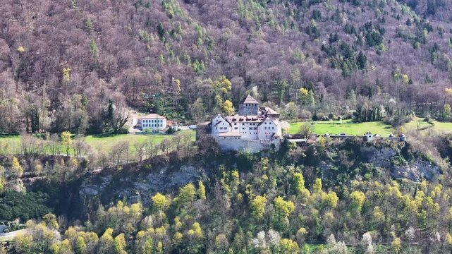 vaduz castle hilltop fortress liechtenstein capital medieval fortification white walls red tile rooftops perched rocky cliff forested hillside spring foliage green valley meadows drone aerial footage
