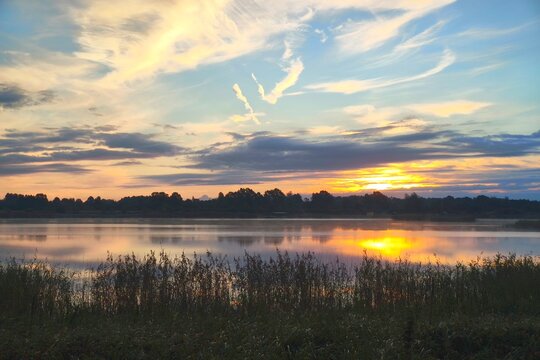 Scenic sunrise over a quiet lake with sun glare on the water surface, reeds (Phragmites australis), and colorful dawn sky.
