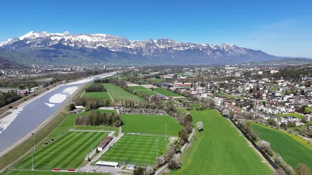town rooftops rhine valley aerial drone footage residential buildings tree lined streets liechtenstein switzerland border green farmland sports pitches river gravel bed alpine mountains landscape