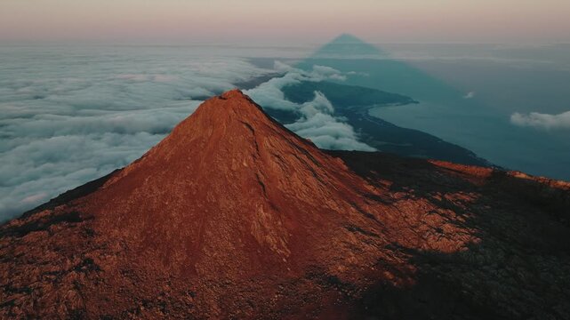 Majestic aerial perspective of the Mount Pico volcano summit at sunset, highlighting its perfect triangular shadow cast over the Atlantic cloud layer and the island's coastline.