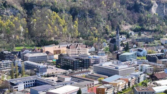 government house town hall vaduz civic center aerial orbit st florin cathedral spire modern office building forested hillside exposed rock face backdrop liechtenstein drone footage heritage site