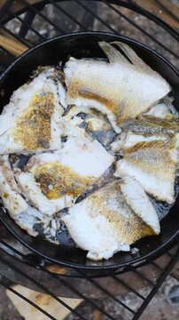 Whole fish portions cook in circular pan over wire grate, with silver skin and spice speckles visible. Heat from charcoal gives surface roasted look and home style texture over hot embers.
