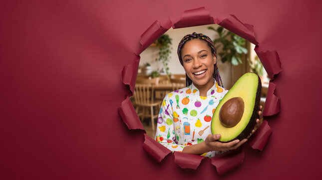 Smiling chef holding a large avocado half through torn paper hole, colorful kitchen uniform. Food service, healthy eating, nutrition, cooking concept.