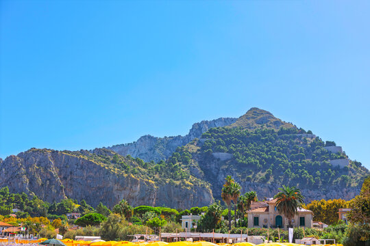 Mondello Beach in Palermo, Italy, sandy shoreline lined with bright sun umbrellas. Towering tree-covered limestone mountain rises into a blue sky behind elegant coastal villas in Sicily
