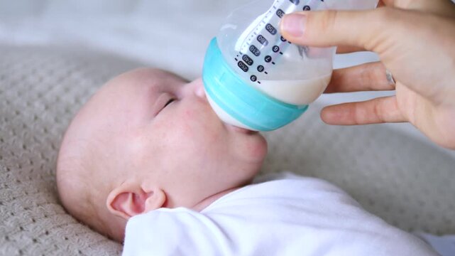 Baby drinks from bottle while lying on soft blanket, calm infant feeding in close up. Adult hand guides milk bottle to baby mouth during gentle nursery care, with newborn resting on bed.