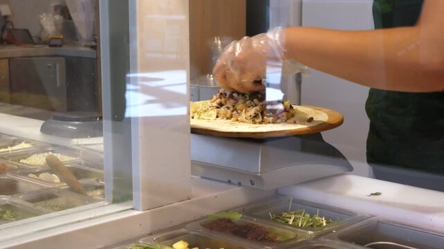 Cafeteria worker finishes pizza assembly with gloved hands, adding toppings across flatbread in fast moving service area. Focused food service employee stands beside ingredient trays and warm