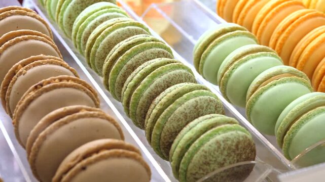 Bakery macarons move along display trays, each almond cookie stacked in tidy rows with creamy beige, green, and orange shells. Sweet macaron assortment looks cheerful and elegant inside dessert shop.