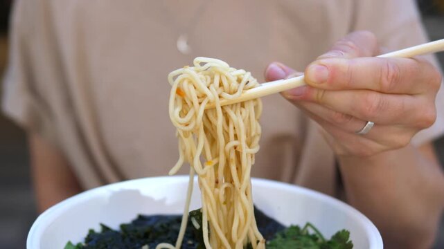 Adult diner lifts noodles with chopsticks from bowl, focused and hungry in casual dining style. Adult diner twirls long strands above vegetable broth, eating at table with steady hands and simple