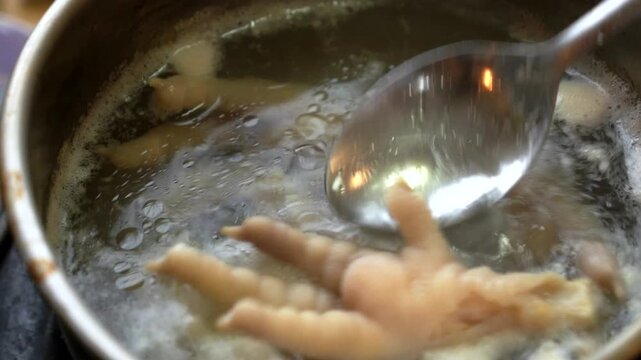 Chicken feet simmer in pot while spoon stirs broth, with boiling cooking process and glossy skin turning tender. Chicken feet remain centered in hot liquid as bubbles rise and cooking continues.