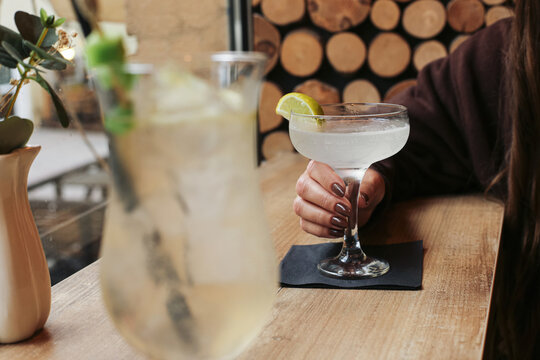 Woman with pale cocktail in coupe glass background. Lime wedge black napkin and tall blurred drink on wooden table. Alcohol beverage refreshment social drinking leisure nightlife and bar lifestyle.