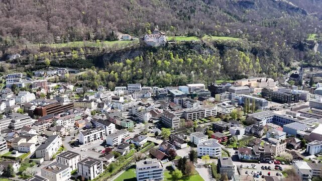 vaduz castle close aerial push in round tower stone walls exposed rock face cliff edge forested hillside town center modern buildings rooftops below drone footage elevated view liechtenstein