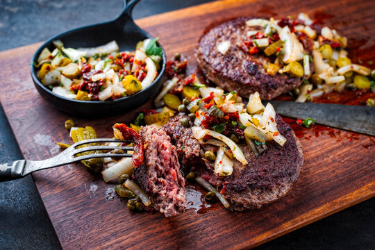 Grilled beef hamburger patties with relish made of gherkin, onions and sun dried tomatos served as close-up on a rustic wooden board