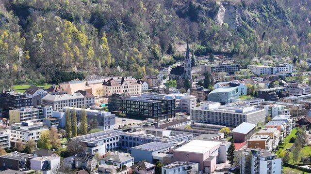 civic district vaduz town center historic buildings cluster st florin cathedral spire modern black facade office building rock face cliff backdrop liechtenstein aerial view drone footage heritage site