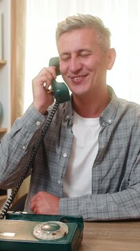Middle-aged man on home table gets pleasant call on retro vintage phone and smiles with joy indoors. Young adult boy on home table laughs during friendly talk and enjoys warm nostalgic communication.