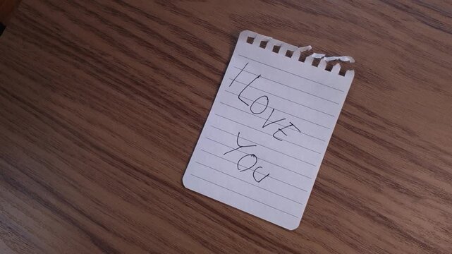 A male hand gently sweeps away a handwritten note reading I Love You to uncover a secret marriage proposal asking Will You Marry Me resting on a wooden table.