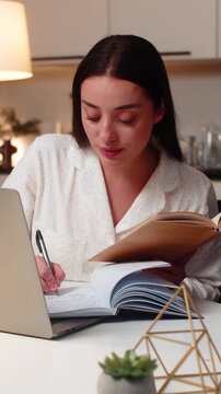 Young woman at night home studies on laptop watches lesson takes notes solves test prepares final exams. Caucasian girl on sofa at evening keeps book near writes outline reviews theory calm study time