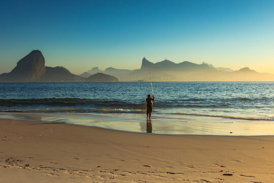 A man fishes at sunset on a beach in Niter&oacute;i, overlooking the waters of Guanabara Bay and iconic Rio de Janeiro landmarks&mdash;Morro da Urca and Christ the Redeemer&mdash;in the background. Brazil.