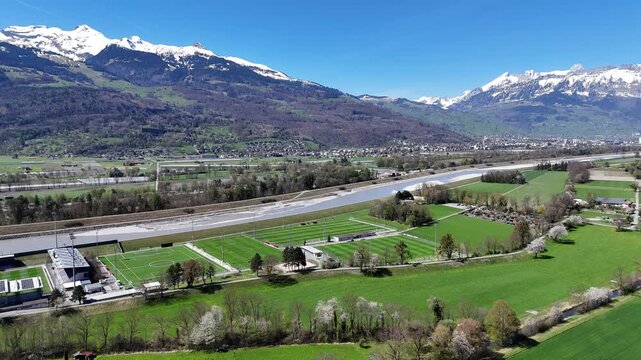running track football pitch sports complex aerial pan rhine river gravel bed snow capped alpine peaks green farmland liechtenstein switzerland border vaduz vineyard hillside distant town rooftops
