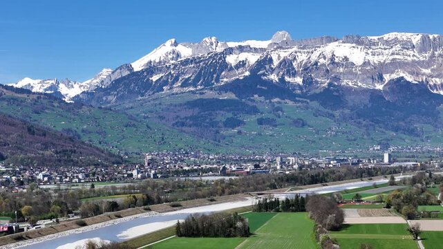 massive alpine ridge snow capped peaks towering rhine valley aerial view distant town rooftops gravel riverbed green farmland foreground liechtenstein switzerland border drone footage panorama