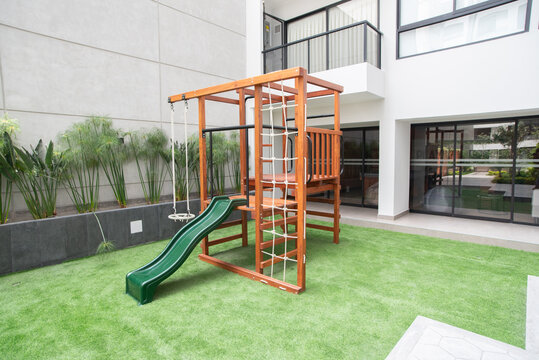Wooden Playground Set: A vibrant wooden playground structure featuring a slide and swing set sits on a soft, artificial grass lawn, bordered by modern architecture and lush greenery.