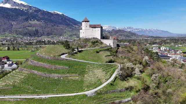 vineyard terraces steep hillside gutenberg castle stone tower turret drone aerial orbit alpine peaks mountain range green farmland valley town rooftops liechtenstein rhine valley medieval fortress