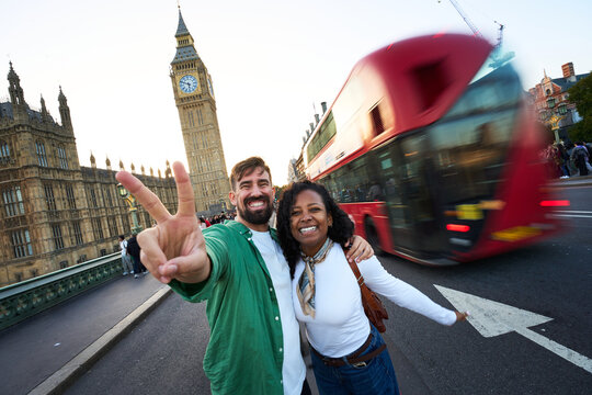 Happy interracial couple posing on Westminster Bridge with Big Ben and a moving red double-decker bus behind. Wide-angle travel portrait conveying excitement, friendship and London energy.